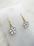 Pair of Kawaii enamel white flower dangle earrings featuring five-petal white blossoms with gold centers and gold-tone outlines, displayed on a marble background.