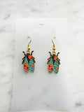 Pair of Kawaii enamel cicada dangle earrings featuring turquoise wings with red floral detailing, outlined in gold-tone metal with gold ear hooks, displayed on a marble background.