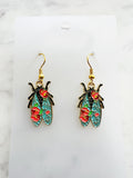 Pair of Kawaii enamel cicada dangle earrings featuring turquoise wings with red floral detailing, outlined in gold-tone metal with gold ear hooks, displayed on a marble background.