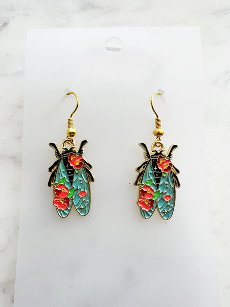 Pair of Kawaii enamel cicada dangle earrings featuring turquoise wings with red floral detailing, outlined in gold-tone metal with gold ear hooks, displayed on a marble background.