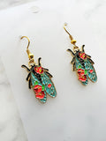 Side view of a Pair of Kawaii enamel cicada dangle earrings featuring turquoise wings with red floral detailing, outlined in gold-tone metal with gold ear hooks, displayed on a marble background.