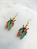 Side view of a Pair of Kawaii enamel cicada dangle earrings featuring turquoise wings with red floral detailing, outlined in gold-tone metal with gold ear hooks, displayed on a marble background.