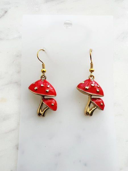Pair of Kawaii red enamel mushroom dangle earrings featuring glossy red caps with raised gold dots and gold-tone stems, displayed on a marble background.