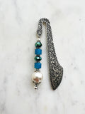 Handmade antique silver bookmark with ornate etched design, featuring blue cube beads, a faceted teal bead, and a pearl accent, displayed on a marble background.