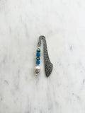 Handmade antique silver bookmark with ornate etched design, featuring blue cube beads, a faceted teal bead, and a pearl accent, displayed on a marble background.