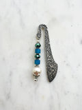 Handmade antique silver bookmark with ornate etched design, featuring blue cube beads, a faceted teal bead, and a pearl accent, displayed on a marble background.
