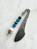 Side view of a Handmade antique silver bookmark with ornate etched design, featuring blue cube beads, a faceted teal bead, and a pearl accent, displayed on a marble background.