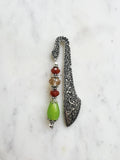 Handmade antique silver bookmark with ornate etched pattern, featuring Tibetan silver bead caps and beads in amber, red, and a lime-green teardrop, displayed on a marble background.