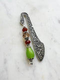 Side view of a Handmade antique silver bookmark with ornate etched pattern, featuring Tibetan silver bead caps and beads in amber, red, and a lime-green teardrop, displayed on a marble background.