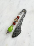 Side view of a Handmade antique silver bookmark with ornate etched pattern, featuring Tibetan silver bead caps and beads in amber, red, and a lime-green teardrop, displayed on a marble background.