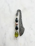 Handmade antique silver bookmark with ornate etched pattern, featuring black faceted beads, a mauve pearl bead, and an olive-green faceted teardrop with Tibetan silver bead caps, displayed on a marble background.