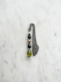 Handmade antique silver bookmark with ornate etched pattern, featuring black faceted beads, a mauve pearl bead, and an olive-green faceted teardrop with Tibetan silver bead caps, displayed on a marble background.