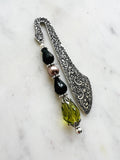 Side view of a Handmade antique silver bookmark with ornate etched pattern, featuring black faceted beads, a mauve pearl bead, and an olive-green faceted teardrop with Tibetan silver bead caps, displayed on a marble background.