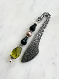 Side view of a Handmade antique silver bookmark with ornate etched pattern, featuring black faceted beads, a mauve pearl bead, and an olive-green faceted teardrop with Tibetan silver bead caps, displayed on a marble background.