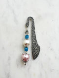 Handmade antique silver bookmark with ornate etched design, featuring blue cube beads, a faceted bead, and a pearl accent, displayed on a marble background.