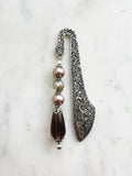Handmade antique silver bookmark with ornate etched pattern, featuring mauve glass pearl beads, a champagne crystal bead, and a glass plum teardrop bead with Tibetan silver bead caps, displayed on a marble background.