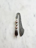 Handmade antique silver bookmark with ornate etched pattern, featuring mauve glass pearl beads, a champagne crystal bead, and a glass plum teardrop bead with Tibetan silver bead caps, displayed on a marble background.