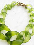 Chunky frosted acrylic chain necklace in translucent green tones, with large twisted links and a polished gold-tone clasp, displayed on a white marble background.