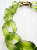 Chunky frosted acrylic chain necklace in translucent green tones, with large twisted links and a polished gold-tone clasp, displayed on a white marble background.