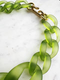 Chunky frosted acrylic chain necklace in translucent green tones, with large twisted links and a polished gold-tone clasp, displayed on a white marble background.