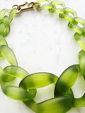 Chunky frosted acrylic chain necklace in translucent green tones, with large twisted links and a polished gold-tone clasp, displayed on a white marble background.