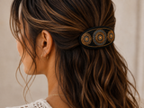 A close-up side view of a woman with long wavy brown hair wearing a black hair clip decorated with gold and orange circular beadwork, set against a soft beige background.