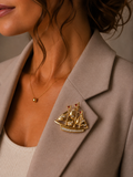 A gold ship-shaped brooch with clear crystal details pinned to the lapel of a taupe blazer worn by a model.