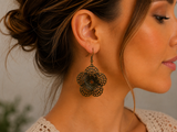 A close-up side profile of a woman wearing a single dangling flower-shaped earring in an antique bronze metal finish.