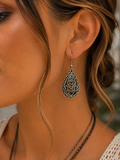 A close-up of a woman's ear wearing a silver-toned teardrop dangle earring with intricate filigree cutouts and scrollwork.