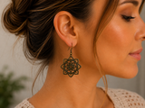 A close-up side profile of a woman wearing a dangling geometric earring with an intricate circular mandala-like pattern in antique bronze.