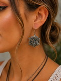 A close-up of a woman's ear wearing a dangling ornate silver-toned earring with an intricate floral or snowflake-like filigree design.