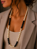 A close-up of a woman wearing a double-strand pearl necklace with alternating strands of white pearls and glossy black beads, styled with a beige blazer and ribbed top.