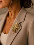 A close-up of a woman wearing a gold fleur-de-lis brooch pinned to her beige blazer.