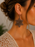 A close-up of a woman wearing a dark bronze filigree flower-shaped dangle earring, showing the intricate metalwork against her neck and ear.