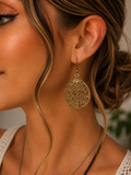 A close-up of a woman wearing a large, ornate gold teardrop-shaped dangle earring with intricate filigree detailing.
