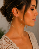 A close-up side profile of a woman wearing a pair of large, thin gold hoop earrings, paired with a beige knit top against a neutral gray background.