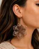 A close-up of a woman wearing a large, ornate filigree dangle earring in a warm metallic bronze color.