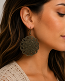 A close-up of a woman wearing a large, ornate filigree dangle earring in an antique bronze finish.