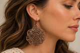 A close-up side profile of a woman wearing a large, ornate bronze filigree dangle earring.
