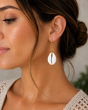 A close-up of a woman wearing a single white cowrie shell dangle earring with gold-tone beads and a central brown textured stripe, shown against a soft neutral background.