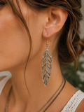 A close-up of a woman's ear wearing a silver-toned dangling feather earring, with her neck and hair visible against a softly blurred background.
