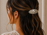 A close-up side profile of a woman with long, wavy dark brown hair wearing a silver-toned metal hair clip adorned with black script engraving. She is wearing a white crochet top and a small gold hoop earring against a soft beige background.