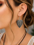 A close-up of a woman's ear wearing a textured, antiqued silver leaf-shaped dangle earring, with her hair in a low bun and layered necklaces visible.