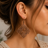 A close-up of a woman wearing a large, ornate copper-toned dangle earring with intricate scrollwork and a textured center, shown against her ear and neck.