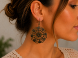 A close-up side profile of a woman wearing a large, intricate black filigree dangle earring. The earring features a detailed circular lace-like pattern hanging from a dark metal hook.