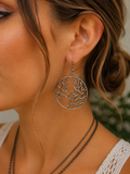 A close-up of a woman's ear wearing a silver tree-of-life dangle earring, with her hair in a low bun and layered necklaces visible against a softly blurred background.