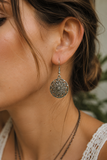 A close-up lifestyle shot of a woman wearing a silver filigree dangle earring. The earring features an intricate circular tree of life design.