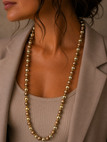 A close-up of a model wearing a long beaded necklace with alternating metallic gold and matte taupe beads, styled with a beige top and blazer.