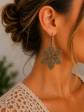 A close-up lifestyle shot of a woman wearing a dangling flower-shaped earring. The earring is made of an antique bronze metal with intricate openwork detailing.