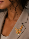 A close-up lifestyle shot of a woman wearing a gold butterfly brooch with orange enamel and a red heart-shaped center, pinned to her taupe blazer.